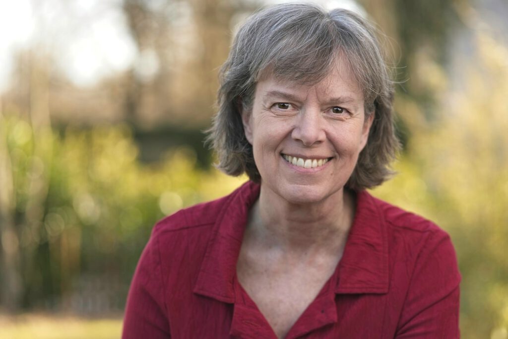 Angelika Neumann Die Künstlerin, Angelika Neumann, in einer roten Bluse mit grünem Garten im Hintergrund