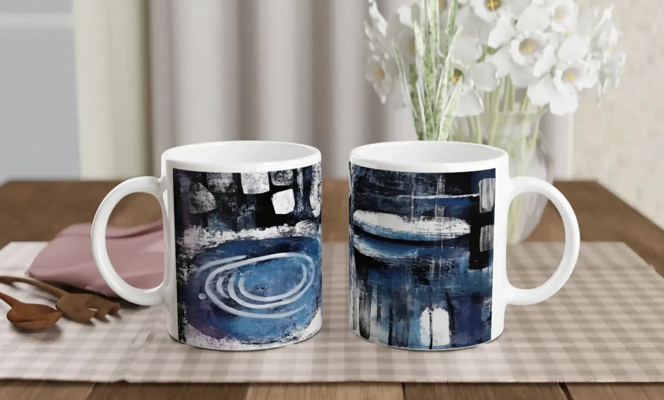 Bildschirmfoto 2025-07-07 um 17.07.16 Zwei weiße Keramikbecher mit abstrakten blauen, schwarzen und weißen Motiven stehen auf einem karierten Tischset auf einem Holztisch. Im Hintergrund sind eine Vase mit weißen Blumen und Utensilien zu sehen.