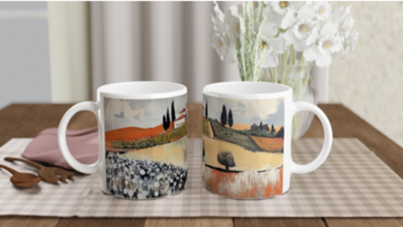Zwei weiße Keramikbecher mit Landschaftsbildern von Feldern, Bäumen und Hügeln stehen nebeneinander auf einem karierten Tischset. Im Hintergrund befinden sich weiße Blumen, Geschirr und eine gefaltete Serviette.