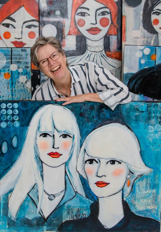 AN-123-E Kopie Eine lächelnde Person mit Brille und gestreiftem Hemd steht hinter einem großen Gemälde zweier Frauen mit hellem Haar, umgeben von anderen farbenfrohen Porträts von Frauen mit roten Lippen und rosigen Wangen.